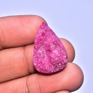 Könnte beinhalten: Ein tropfenförmiger, grob strukturierter, fuchsiafarbener Edelstein. Der Stein hat ein Loch in der Nähe der Oberseite. Der Edelstein wird in einer Hand vor einem weißen Hintergrund gehalten. Die Oberfläche des Steins hat ein körniges Aussehen.