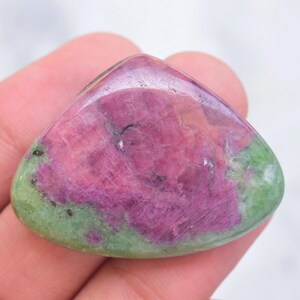 May include: A polished, triangular-shaped ruby zoisite gemstone featuring a vibrant magenta-pink center surrounded by a smooth, light green border with small black mineral inclusions throughout the surface of the stone.