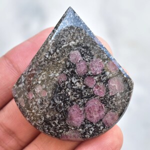 Peut inclure: Une pierre précieuse polie avec un fond sombre moucheté et des cristaux rose-rouge incrustés. La pierre a une forme arrondie en forme de bouclier avec une surface brillante et réfléchissante. Les cristaux sont de forme irrégulière et de tailles variées.