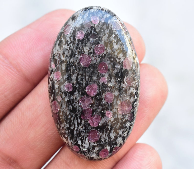 K&ouml;nnte beinhalten: Ein ovaler Cabochon-Edelstein mit einer polierten Oberfl&auml;che und einer schwarz-wei&szlig; gesprenkelten Matrix. Der Stein ist mit zahlreichen kleinen, durchscheinenden, kreisf&ouml;rmigen und unregelm&auml;&szlig;igen magentafarbenen Einschl&uuml;ssen durchsetzt.