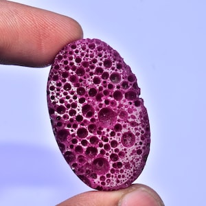 Könnte beinhalten: Ein poröses, oval geformtes Objekt in tiefem Magenta. Die Oberfläche ist mit zahlreichen Löchern unterschiedlicher Größe bedeckt. Gehalten zwischen Fingern vor hellblauem Hintergrund.