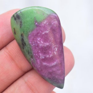 May include: A polished, teardrop-shaped zoisite gemstone featuring a vibrant magenta-pink center surrounded by opaque lime-green mineral deposits and small, dark black inclusions throughout the smooth, reflective surface of the stone.