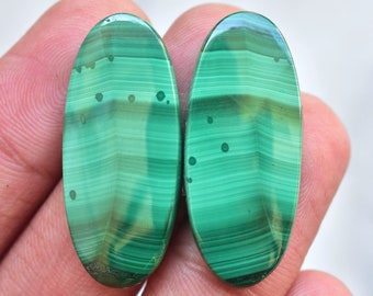 Puede incluir: Dos piedras ovaladas pulidas de malaquita. Las piedras son de un verde vibrante con patrones de bandas verdes y marrones más oscuros. Pequeños puntos oscuros están dispersos por la superficie. Las piedras tienen un acabado liso y brillante.