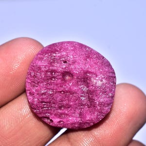 May include: A rough, round, deep pink ruby gemstone. The surface has a textured, granular appearance with small pits and variations in color. The gemstone is held in a hand against a light background.