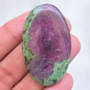 May include: An oval-shaped polished gemstone featuring a vibrant magenta-purple center surrounded by mottled light green and dark green mineral inclusions. The stone has a smooth, glossy surface texture with visible natural crystalline patterns.