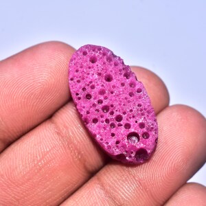 Könnte beinhalten: Ein lebhaft magentafarbener, poröser Stein mit unregelmäßiger ovaler Form. Der Stein hat zahlreiche kleine und große Löcher auf seiner Oberfläche, wodurch ein strukturiertes Aussehen entsteht. Der Stein wird in einer Hand vor weißem Hintergrund gehalten.