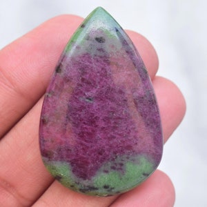 May include: A polished, pear-shaped ruby zoisite gemstone featuring a vibrant magenta-pink center surrounded by mottled lime green areas and scattered black mineral inclusions throughout the smooth, rounded surface of the stone.