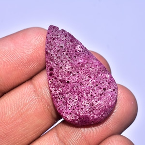 Può includere: Una gemma grezza a forma di goccia, di colore magenta intenso. La pietra ha una consistenza porosa ed è tenuta in mano su uno sfondo chiaro. Questa pietra naturale potrebbe essere utilizzata per la creazione di gioielli o come elemento decorativo.