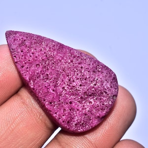 Könnte beinhalten: Ein lebendiger, unregelmäßig geformter, magentafarbener Edelstein mit poröser Textur. Der Stein wird in einer Hand vor einem hellblauen Hintergrund gehalten. Dieser einzigartige Stein könnte für Schmuck oder zur Dekoration verwendet werden.