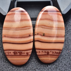 May include: Two polished oval cabochons featuring horizontal bands of light peach, tan, and dark brown colors. The stones display a smooth, glossy surface with distinct layered patterns and small, dark, moss-like inclusions near the bottom.