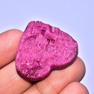 Könnte beinhalten: Ein grob geschnittener, herzförmiger Edelstein in leuchtendem Magenta. Der Stein hat eine strukturierte Oberfläche mit sichtbaren Kristallstrukturen und kleinen Hohlräumen. Der Edelstein wird in einer Hand vor weißem Hintergrund gehalten.