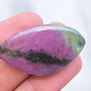 May include: A marquise-shaped polished gemstone featuring a vibrant magenta-pink center with a prominent dark charcoal-black horizontal band and a patch of mossy green at the right edge, showcasing a smooth, reflective surface and natural mineral inclusions.