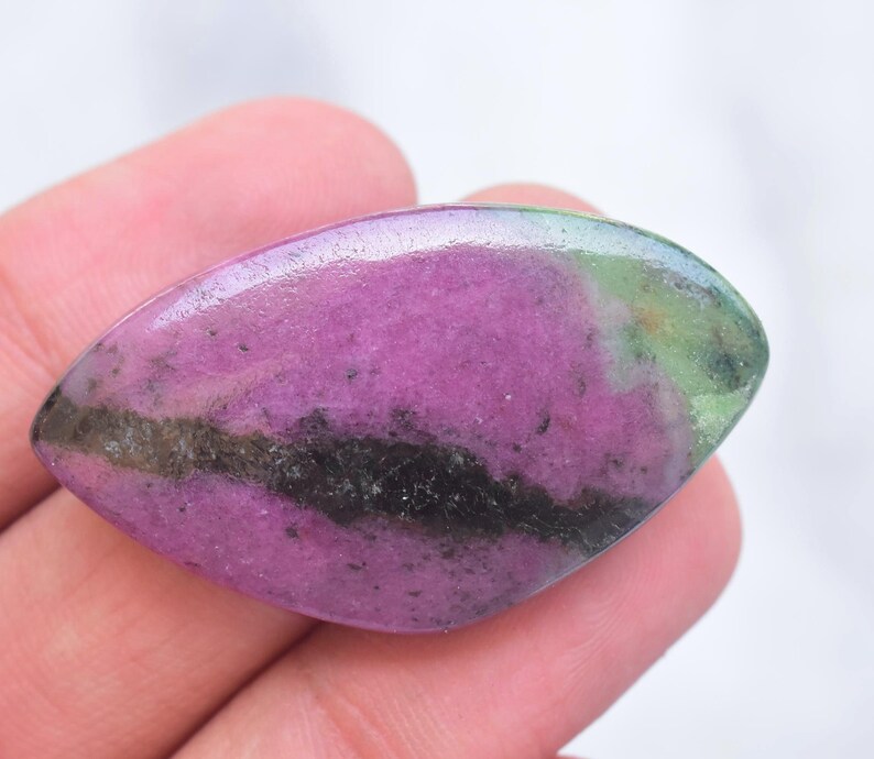 May include: A marquise-shaped polished gemstone featuring a vibrant magenta-pink center with a prominent dark charcoal-black horizontal band and a patch of mossy green at the right edge, showcasing a smooth, reflective surface and natural mineral inclusions.