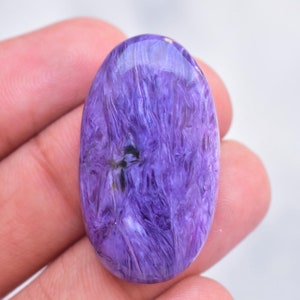 Op de afbeelding: Een ovaalvormige cabochon edelsteen met een levendige paarse kleur, voorzien van wervelende, vezelige patronen en donkere insluitingen. De steen heeft een glad, gepolijst oppervlak dat licht reflecteert en de natuurlijke textuur toont.
