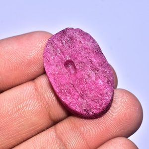 Könnte beinhalten: Ein grob geschnittener, oval geformter Rubin-Edelstein mit tiefrosa Farbton. Der Stein hat eine natürliche, strukturierte Oberfläche und ein kleines, gebohrtes Loch. Der Edelstein wird in einer Hand vor hellem Hintergrund gehalten.