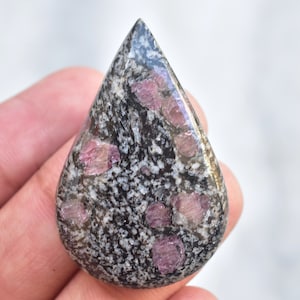 May include: A polished, teardrop-shaped gemstone. The stone has a speckled pattern of black, grey, and white, with several vibrant pink inclusions. The gem is held in a hand.