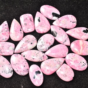 Puede incluir: Una colección de piedras preciosas de rodonita pulidas en varias formas, incluyendo lágrimas y óvalos. Las piedras muestran un vibrante tono rosa con inclusiones negras y grises, sobre un fondo oscuro.