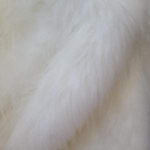 May include: White faux fur fabric with a soft, fluffy texture. The fur is arranged in a diagonal pattern, creating a sense of movement and depth.