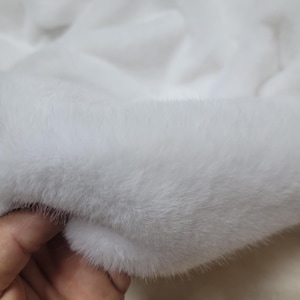 Puede incluir: Primer plano de una tela de piel sintética blanca y suave. La textura es densa y afelpada, con fibras largas y fluidas. La tela es de color blanco liso, con sutiles variaciones de luz y sombra.