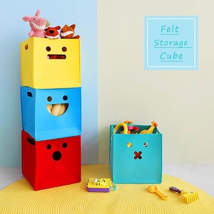 Peut inclure: Trois cubes de rangement en feutre colorés empilés, chacun avec un design de visage différent. Le cube supérieur est jaune, le milieu est bleu et le bas est rouge. Un quatrième cube turquoise est à côté de la pile. Des jouets sont à l'intérieur et sur les cubes. Le texte "Felt Storage Cube" est dans un encadré bleu.