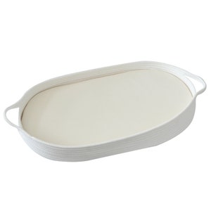 May include: An oval-shaped white woven tray with a cream-colored interior. The tray has two curved handles and is made of a thick, textured rope-like material. The interior is smooth and flat.