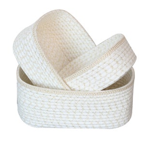 Puede incluir: Dos cestas de almacenamiento tejidas blancas con detalles dorados. Las cestas son de forma ovalada y están apiladas una encima de la otra.
