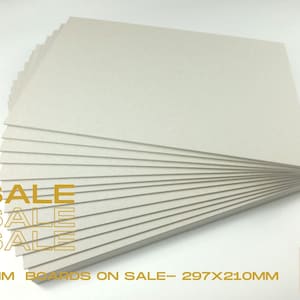 May include: A stack of 3mm thick grey cardboard sheets, each measuring 297 x 210 mm. The text "SALE" is printed in yellow on the bottom left corner of the image. The text "3MM BOARDS ON SALE - 297X210MM" is printed in yellow on the bottom of the image. The text "TBB TRADITIONAL BOOK BINDING" is printed in black on a white background in the bottom right corner of the image.
