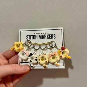 May include: A set of handmade stitch markers featuring cat-shaped designs. The markers are attached to metal rings and displayed on a white card with the text "HANDMADE STITCH MARKERS". The cat markers have various colors, including yellow, white, black, and pink.