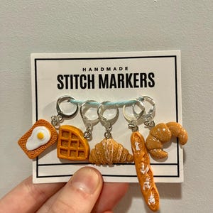May include: Handmade stitch markers with a breakfast theme. The markers feature a fried egg, waffle, croissant, and baguette.
