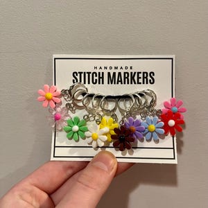 Puede incluir: Un conjunto de marcadores de punto hechos a mano con dijes de flores de margarita de colores. Los marcadores de punto están unidos a anillos de color plateado y se muestran en una tarjeta blanca con texto negro que dice "HANDMADE STITCH MARKERS". Las flores son de varios colores, incluyendo rosa, verde, amarillo y morado.