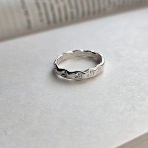 Op de afbeelding: Een zilveren ring met een golvend ontwerp en kleine hartvormige uitsparingen. De ring ligt op een wit oppervlak, mogelijk een pagina uit een boek, met de randen van het boek zichtbaar op de achtergrond. De ring lijkt van glanzend metaal te zijn.