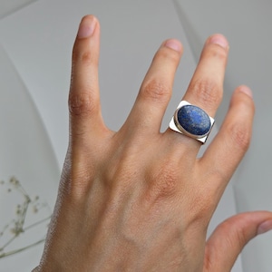 Peut inclure: Une bague en argent ornée d'une grande pierre précieuse lapis-lazuli de forme ovale, d'un bleu profond avec de subtiles taches dorées. La pierre est sertie dans une monture en argent lisse et poli sur un anneau large, plat et rectangulaire.