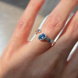 Peut inclure: Une bague ouverte de couleur argentée ornée d'une pierre précieuse bleue de forme ovale sertie dans un chaton lisse. La bague présente une finition métallique polie et une ouverture sur le dessus, avec un design simple et épuré.