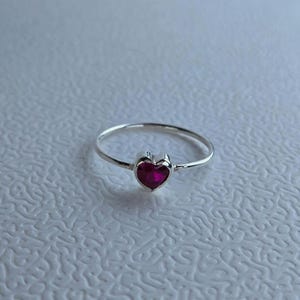 Peut inclure: Une bague en argent avec une pierre précieuse rouge foncé en forme de cœur. La monture de la bague est fine et polie, et la pierre en forme de cœur est sertie dans une lunette simple et élégante. La bague est présentée sur une surface blanche texturée.