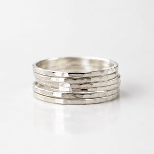 Peut inclure: Une pile de cinq bagues fines en argent, chacune présentant une surface martelée et texturée qui crée une finition métallique réfléchissante. Les bagues sont uniformes en forme et en taille, disposées verticalement pour former un anneau.