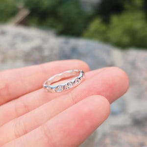 Può includere: Un anello color argento caratterizzato da un design a fascia continua con forme a cuore in rilievo lungo tutta la circonferenza. La superficie metallica presenta una finitura lucida e riflettente che cattura la luce, creando un aspetto liscio.