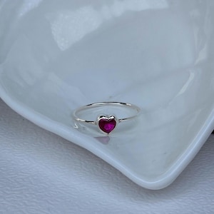 Peut inclure: Une bague en argent avec une pierre précieuse rose foncé en forme de cœur. La bague est sertie sur un fin anneau et repose sur un plat blanc en forme de cœur.