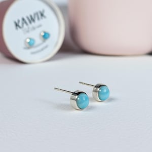 Peut inclure: Deux boucles d'oreilles rondes bleu clair avec des montures en métal argenté. Les boucles d'oreilles sont posées sur une surface blanche, avec une boîte cylindrique et un bol rose en arrière-plan. La boîte porte l'inscription "KAWIK".