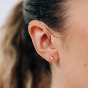 Peut inclure: Gros plan sur une oreille ornée d'une petite boucle d'oreille en forme d'éclair de couleur or. La boucle d'oreille est de conception minimaliste. La peau est claire, les cheveux sont attachés.