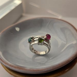 Peut inclure: Une bague en argent composée de trois anneaux distincts qui convergent vers une tige unique. La bague est ornée de trois pierres précieuses rondes taillées en cabochon, aux teintes bleu sarcelle, vert olive et magenta, serties sur les anneaux.