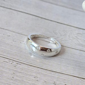 Peut inclure: Une bague en argent poli avec un anneau arrondi, ornée de plusieurs petites pierres précieuses transparentes. La bague est posée sur une surface en bois clair, mettant en valeur son éclat.