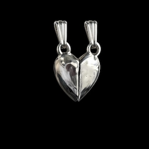 Peut inclure: Deux pendentifs en forme de cœur argentés, chacun représentant la moitié d'un cœur brisé, reliés par une petite boucle en haut. Les pendentifs sont très polis, reflétant la lumière sur un fond noir. Un bijou romantique.
