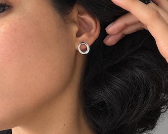 May include: A small, circular stud earring with a silver-toned finish and a textured, hammered surface. The earring is worn in the earlobe, featuring a simple, open-center design that creates a subtle, reflective appearance on the metal surface.