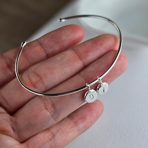 May include: A silver-colored bangle bracelet with two small, round charms. Each charm has a single letter engraved on it. The bracelet is held in a hand, and the background is blurred.