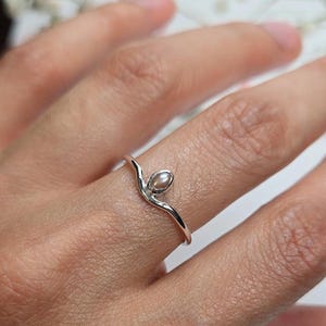 Peut inclure: Une bague en argent délicate ornée d'une petite perle ovale. Le corps de la bague présente un design fluide et organique qui épouse le doigt, la perle étant placée au sommet. La bague est portée sur un doigt.
