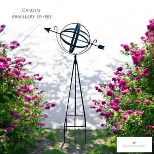 May include: A black metal armillary sphere garden ornament with an arrow, set on a tripod stand. The text "GARDEN ARMILLARY SPHERE" is at the top. The background features pink flowering bushes.