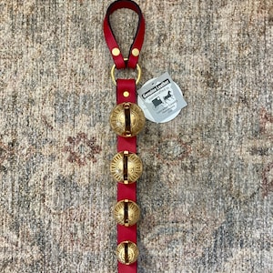 May include: A red leather strap with four brass bells and a loop at the top. The bells are attached to the strap with brass rivets. A tag reads "Genuine Leather Handcrafted in Lancaster County Pennsylvania USA."