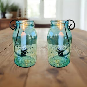 May include: Two teal-colored mason jar candle holders with black metal handles. Each jar holds a lit white candle. The jars are on a wooden table, and the background includes a window and plants.
