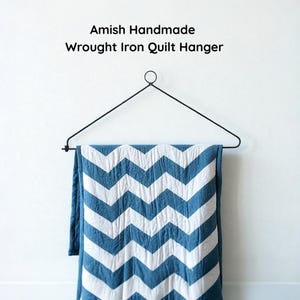 May include: A black wrought iron quilt hanger with a blue and white chevron patterned quilt. The text "Amish Handmade Wrought Iron Quilt Hanger" is above the hanger. The quilt is hanging against a white wall.