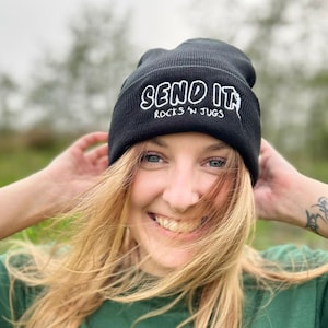 Op de afbeelding: Een vrouw draagt een zwarte beanie met de tekst "SEND IT ROCKS N JUGS" in wit. Ze draagt ook een groen t-shirt met hetzelfde logo in wit.